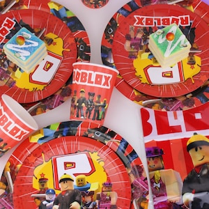Op de afbeelding: Rode feestborden en bekers met Roblox-personages en logo's. De borden hebben een geel en rood ontwerp met een nummer en karakterafbeeldingen. Kleine vierkante cakes met kleurrijke glazuur liggen op de borden. Een rode banner met het Roblox-logo is ook zichtbaar.