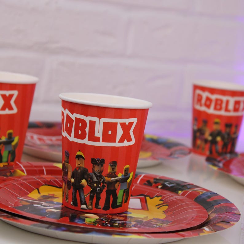 Op de afbeelding: Rode papieren bekers en borden met een Roblox-thema. De bekers en borden hebben het Roblox-logo en karakterillustraties. De bekers hebben een witte rand en zijn rood met verticale strepen.