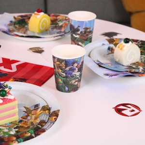 May include: A party scene with themed paper cups, plates, and desserts. The cups and plates feature colorful cartoon characters. The table is set with small cakes and a red and white striped bag.