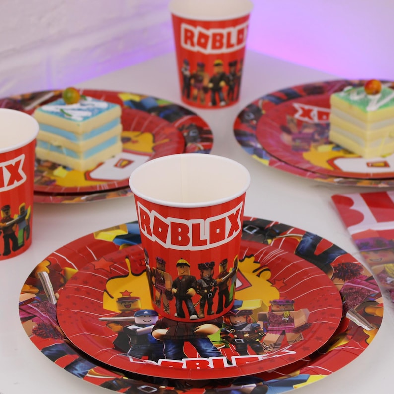 Op de afbeelding: Rode papieren bekers en borden met een Roblox-thema. De bekers en borden tonen het Roblox-logo en karakterillustraties. De borden zijn rood met een ster- en karakterontwerp. Er zijn ook stukjes cake te zien.