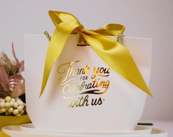May include: White gift bag with a yellow satin ribbon and gold lettering that says "Thank you for celebrating with us."