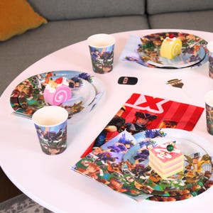 May include: A party table setting with plates, cups, and napkins featuring a colorful cartoon design. The plates hold slices of cake. The table is white and the background includes a gray sofa.