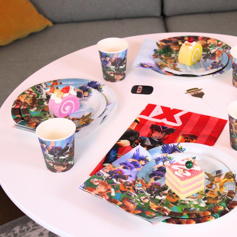 May include: A party table setting with plates, cups, and napkins featuring a colorful cartoon design. The plates hold slices of cake. The table is white and the background includes a gray sofa.