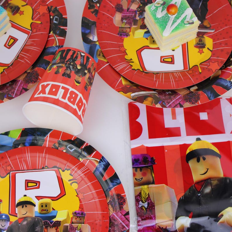 Op de afbeelding: Rode papieren borden en een beker met Roblox-personages en logo. De borden hebben een geel ontwerp met een rode rand. Een klein stukje cake met groene glazuur ligt op een bord. Feestartikelen.
