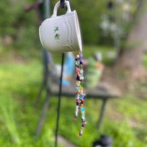 Peut inclure: Une tasse à thé en céramique blanche avec une anse, ornée de motifs de feuilles de lierre vertes, semble verser un flot de perles colorées. La tasse est suspendue à un crochet en métal noir sur un fond vert flou.