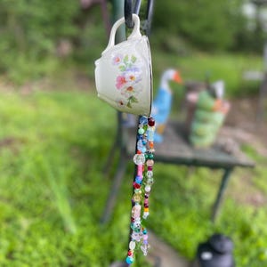 Peut inclure: Une tasse à thé blanche avec un motif floral est suspendue à un crochet en métal noir. Une guirlande de perles colorées, roses, bleues et vertes, pend du bord de la tasse. Les perles sont de formes variées.