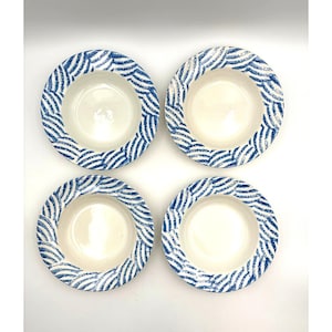 May include: Four white ceramic bowls with a blue wave pattern around the rim. The bowls are round with a shallow depth, and the blue pattern is a repeating design. The bowls are arranged in a square formation against a white background.