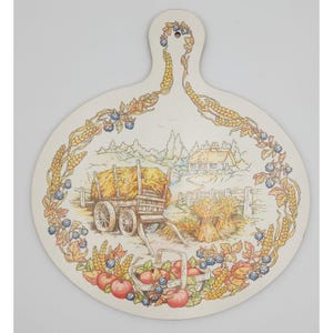 May include: Cream-colored, oval-shaped decorative cutting board with a handle. The board features a pastoral scene with a hay wagon, a cottage, and a basket of apples. Wheat and berries border the scene.