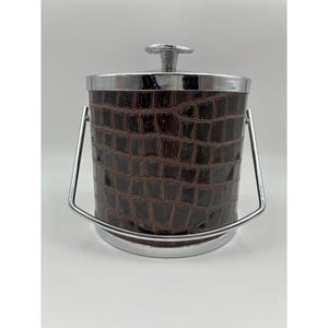 May include: A cylindrical ice bucket featuring a dark brown exterior with a textured crocodile skin pattern. The container is accented with a polished chrome rim, a matching chrome lid with a central knob handle, and a hinged chrome swing handle.