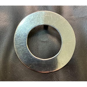 May include: A circular, silver-colored metal object with a hole in the center. The metal has a brushed appearance and shows some wear. The object is set against a black, textured background.