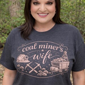 May include: Dark gray t-shirt with a rose gold graphic that reads "coal miner's wife." The design includes a miner's helmet, crossed pickaxes, and a cart filled with coal. The shirt is worn by a person.