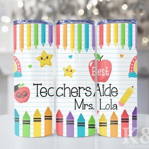 May include: A white tumbler with a school theme. It features colourful pencils, crayons, a red apple with the word "Best", stars, and rainbows. The text "Teachers Aide Mrs. Lola" is also visible.