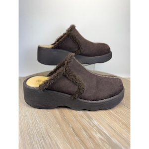 May include: A pair of dark brown platform mules featuring a suede-like upper with dark brown faux fur trim along the collar and a thick, solid dark brown platform sole. The interior footbed is light tan with a circular size indicator visible.