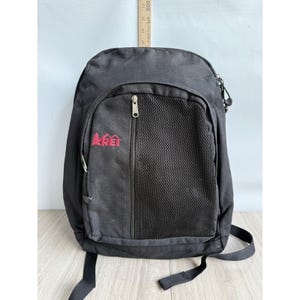 May include: Black backpack with a zippered front pocket featuring a mesh panel. The backpack has a red embroidered logo on the front. Adjustable shoulder straps and a top handle are visible. The backpack is made of a durable, possibly water-resistant material.