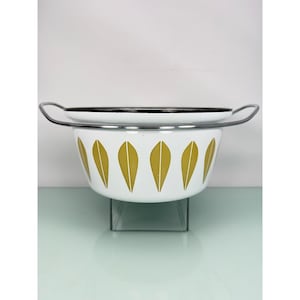 May include: A white enamel colander featuring a repeating pattern of mustard yellow leaf-shaped motifs around the exterior. The bowl has a black rim and two silver-toned metal handles extending from the sides, providing a functional kitchen tool.