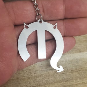 May include: Silver-colored metal keychain with a devil-themed design. The keychain features a stylized letter "T" with horns and a curved tail ending in an arrow. The keychain is attached to a metal ring and chain.