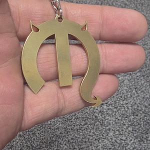 May include: A gold-colored metal keychain featuring a stylized letter M design. The letter has two small pointed horn-like protrusions at the top and a curved, tail-like extension at the bottom right. It is attached to a standard circular metal ring.