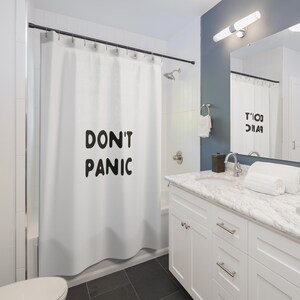 May include: White shower curtain with the words "DON'T PANIC" in bold black letters. The curtain hangs in a modern bathroom with white cabinetry, a marble countertop, and a dark gray tiled floor. A mirror reflects the curtain.