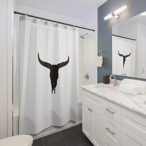 May include: White shower curtain with a black silhouette of a bull skull with large horns. The shower curtain is in a bathroom with a white vanity and a mirror.