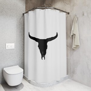 May include: White shower curtain with a black silhouette of a bull skull. The shower curtain is hung on a curved shower rod.