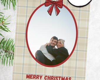 May include: Christmas card with a beige plaid pattern and a red bow illustration. The card has a red oval frame with a photo of a couple. Text at the bottom reads "MERRY CHRISTMAS THE ADAMS | 20XX". A circular button says "CUSTOMIZE WITH CANVA".