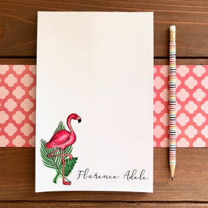 Op de afbeelding: Een wit notitieblok met een roze flamingo-illustratie en de naam "Florence Adele" in script. Een gestreepte potlood ligt naast het notitieblok op een houten oppervlak met een roze patroonrand.