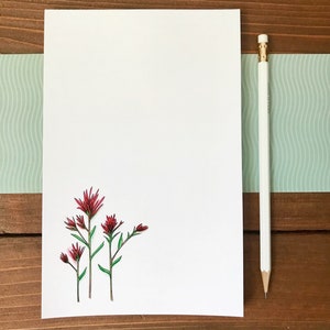 Op de afbeelding: Een wit vel papier met een aquarel illustratie van rode wilde bloemen en groene stelen. Rechts een witte potlood met een gouden bus en gum. De achtergrond is een houten oppervlak.
