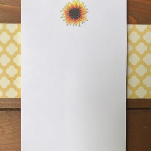 Könnte beinhalten: Weißes Briefpapier mit einer Sonnenblumenillustration oben. Die Sonnenblume hat gelbe Blütenblätter und eine dunkelbraune Mitte. Das Briefpapier ist von gelbem Papier mit weißen Vierpassmustern umrandet. Der Hintergrund ist eine Holzoberfläche.