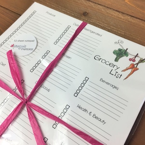 May include: A white grocery list notepad tied with a pink ribbon. The notepad includes sections for produce, dairy, frozen, and other categories. The words "Grocery List" are printed on the top right corner.
