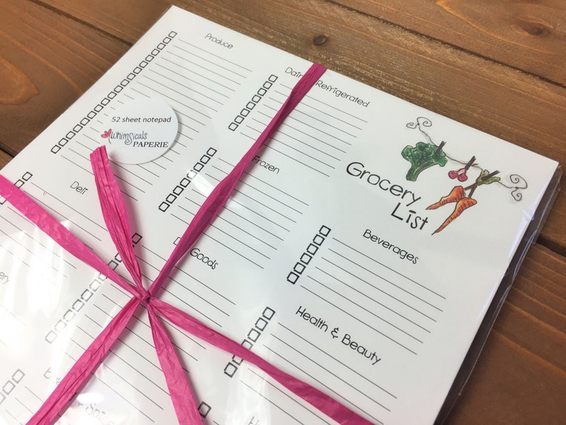 May include: A white grocery list notepad tied with a pink ribbon. The notepad includes sections for produce, dairy, frozen, and other categories. The words "Grocery List" are printed on the top right corner.