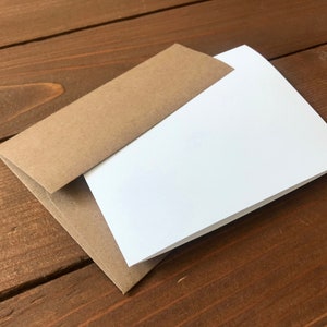 May include: A kraft paper envelope with a white card inside, set against a dark wood grain background. The envelope is open, revealing the card. The scene is lit with natural light, highlighting the textures of the paper and wood.