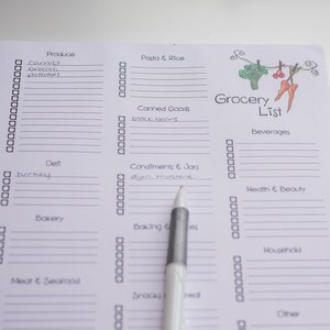 May include: A white grocery list with sections for produce, pasta, canned goods, and more. The list includes handwritten items like carrots, broccoli, and black beans. A pen points to the "Condiments & Jars" section. The words "Grocery List" are at the top.