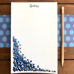 Op de afbeelding: Een witte notitieblok met de naam "Audrey" in zwart schrift bovenaan. De linker benedenhoek is versierd met blauwe en gele hartvormen. Een houten potlood ligt aan de rechterkant van het notitieblok.