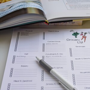 May include: A printed grocery list with check boxes, categories, and the title "Grocery List" in a handwritten font. A pen rests on the list, with an open cookbook and a fork in the background.