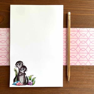 Op de afbeelding: Een blanco wit notitieblok met een potlood en een roze patroonrand. Het notitieblok bevat een aquarelillustratie van twee knuffelende otters, met groene bladeren en schelpen.