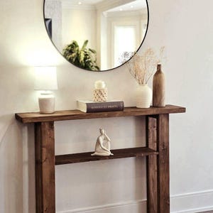 Peut inclure: Une console en bois brun foncé avec une étagère. La table est surmontée d'un miroir rond, et d'une lampe blanche, d'un livre intitulé "Botanical Prints", d'un vase blanc avec des fleurs séchées et d'un vase brun sur le dessus de la table. Une sculpture en céramique blanche est sur l'étagère.