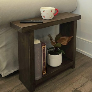 Peut inclure: Une table d'appoint en bois foncé avec une étagère. La table a une tasse en céramique blanche avec des cerises rouges et un nœud rose, une télécommande noire, un livre intitulé "Botany of Desire", un livre rouge et une plante en pot avec des feuilles brunes.