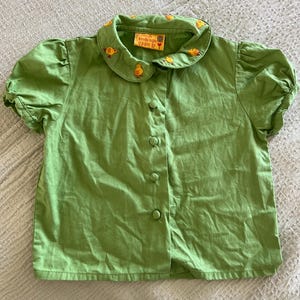 May include: A green button-down shirt with short, puffed sleeves. The collar is adorned with embroidered yellow chicks. The shirt has a tag that reads "breede m d pam 12".