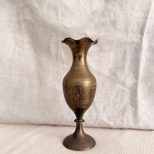 May include: A tall, slender brass vase with a flared, wavy rim and a narrow neck. The body of the vase features horizontal bands of etched, intricate patterns. It stands on a circular pedestal base, showcasing a metallic, aged gold-toned finish.