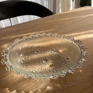 May include: A clear glass decorative plate with a textured surface and a beaded edge. The plate has a circular shape with a central design and is on a wooden surface.