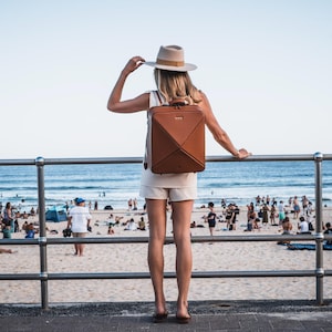 Könnte beinhalten: Eine Frau in einem weißen Hut und weißen Shorts steht auf einer Promenade mit einem braunen Lederrucksack. Die Promenade befindet sich vor einem Strand mit Menschen darauf.