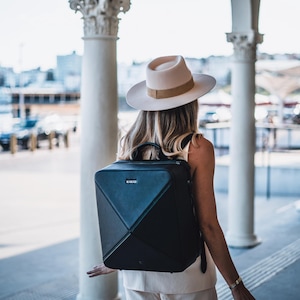 Könnte beinhalten: Ein schwarzer, geometrischer Rucksack mit einem oberen Griff und einem kleinen Logo. Die Person trägt einen cremefarbenen Hut mit einem braunen Band. Der Hintergrund umfasst weiße Säulen und eine verschwommene Außenszene.