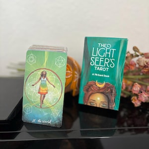 May include: A tarot card deck and box set. The box is teal with the text "THE LIGHT SEER'S TAROT" and an illustration of a woman. The card shows a woman in a rainbow dress with the text "21 the World".