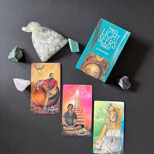May include: The Light Seer's Tarot deck box with a teal cover, three tarot cards titled The Emperor, The Hierophant, and Strength, a sheer white drawstring bag filled with clear crystals, and four loose raw crystals in green, pink, and purple.