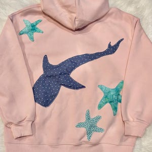May include: Pink hooded sweatshirt with a whale shark applique in navy blue polka dot fabric. Three teal starfish appliques are also on the back of the sweatshirt. The sweatshirt has a ribbed hem and cuffs.