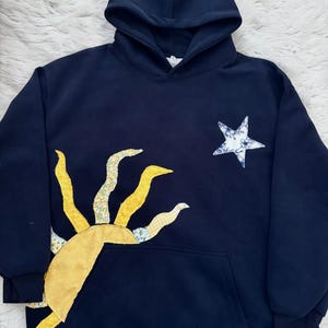 May include: Navy blue hoodie with a large yellow sun and a white and blue floral star applique. The sun has a yellow center and yellow and floral print rays. The hoodie has a front pocket and a hood.