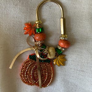 May include: A gold-toned U-shaped metal keychain featuring a beaded pumpkin charm, a thin gold ribbon, and various decorative beads including orange, green, and cream-colored stones, along with small metal leaf charms in orange and yellow.