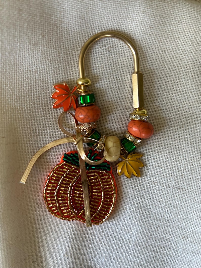 May include: A gold-toned U-shaped metal keychain featuring a beaded pumpkin charm, a thin gold ribbon, and various decorative beads including orange, green, and cream-colored stones, along with small metal leaf charms in orange and yellow.
