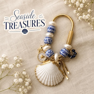 May include: A gold-toned metal keychain featuring a large white seashell, blue and white patterned ceramic beads, white pearls, a small gold heart charm, and a cluster of dark blue beads. The text Seaside Treasures is printed in blue script.