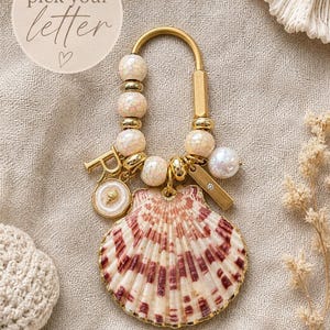 May include: A gold-toned metal keychain featuring a large, fan-shaped seashell with reddish-brown stripes. The hardware includes a U-shaped ring, several cream-colored crackle beads, a gold letter R charm, a small circular shell charm, and a rectangular bar.